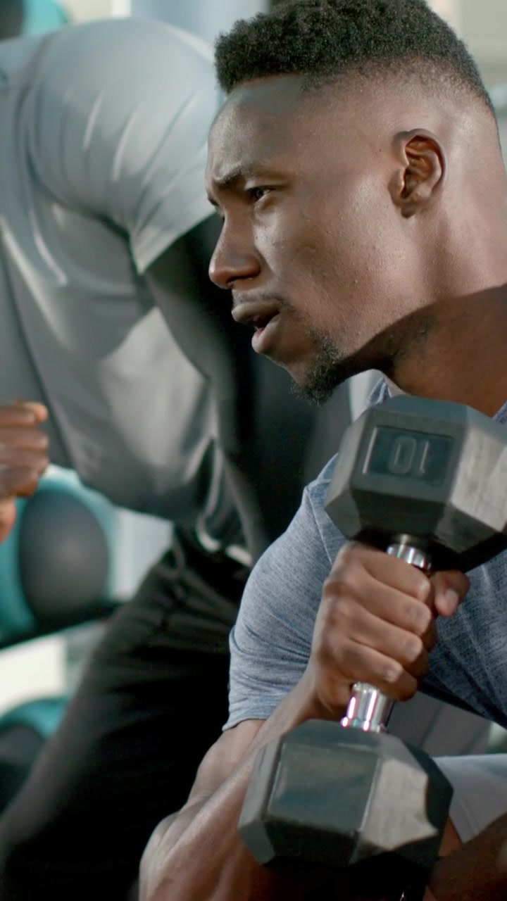 Vertical video: Curling seated trainee on coach cue at gym bench holding 10lb, coach guiding form