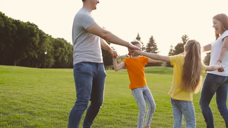 happy parents and little daughters holding hands and spinning around on green grass field in the park