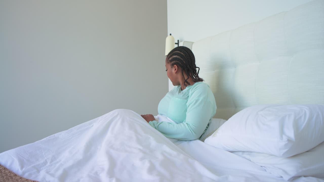Ochtendlicht wekt een vrouw in bed, die rechtop gaat zitten, water drinkt, door haar telefoon scrolt en lacht