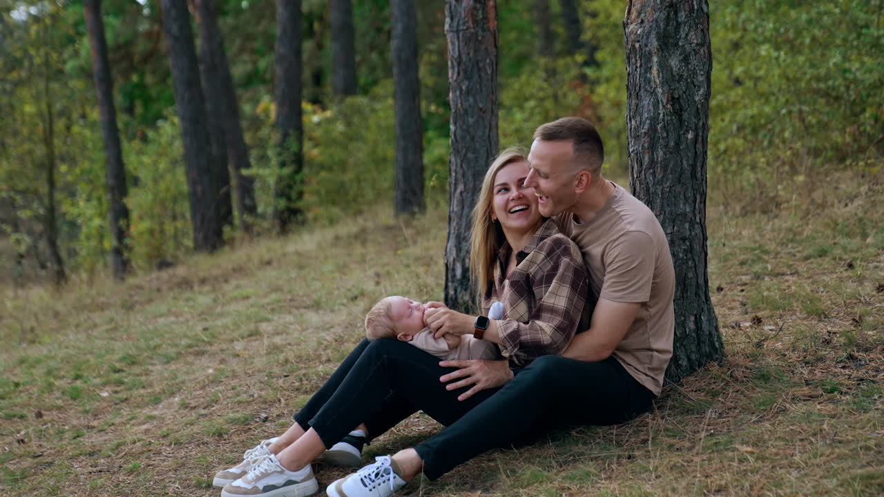 Young parents having good time with their baby in the nature. Woman with child in hands sits leaned on her husband leaning on the tree.