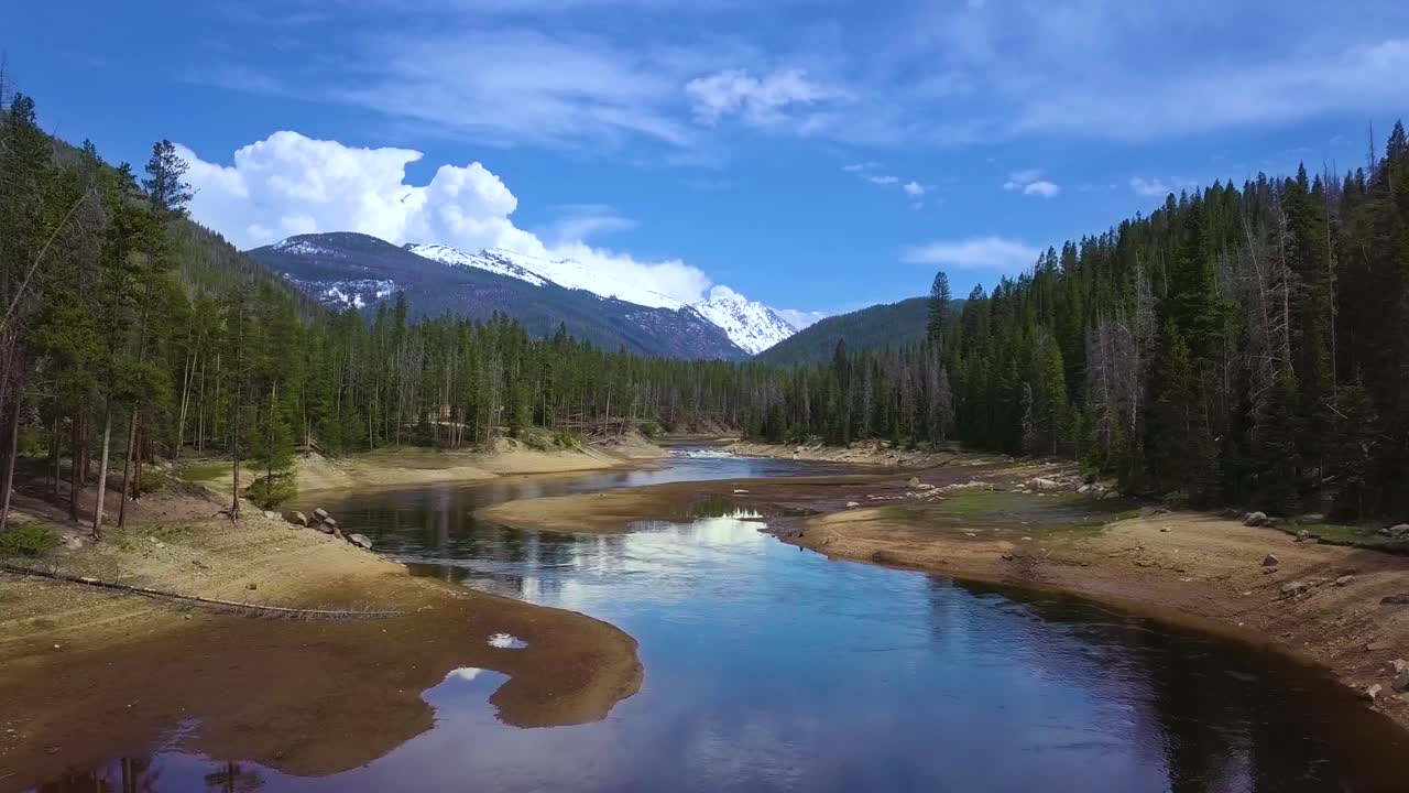 vista aérea baja volando lentamente por un río ancho y sinuoso en la brillante y soleada tarde de primavera en el desierto del bosque con majestuosas montañas cubiertas de nieve, cielo azul y nubes que se reflejan en la superficie del agua