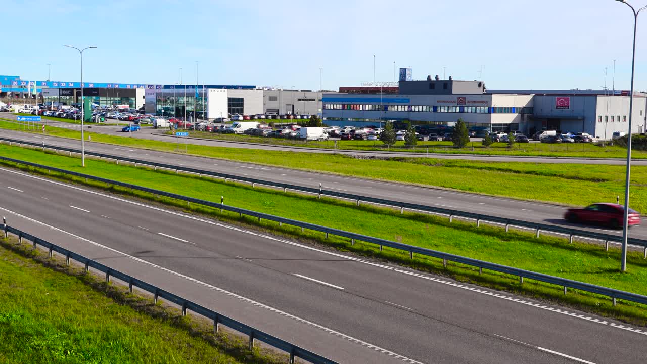 Cars and vehicles driving on an Estonian Pärnu highway during summer time on asphalt with markings and industrial warehouse buildings in the background. Sun is shining and sky is blue, green grass.
