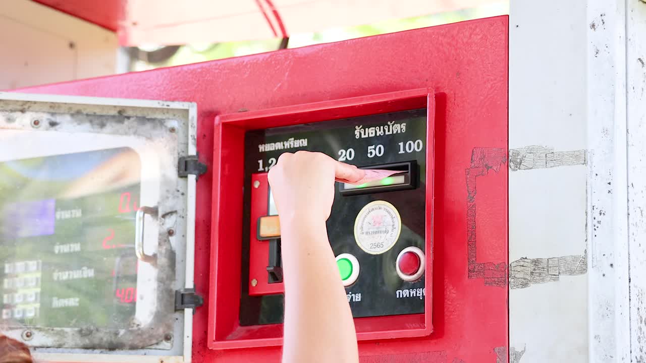A person inserts coins into a red fuel dispenser under bright daylight at Khao Rang Viewpoint, Phuket, Thailand