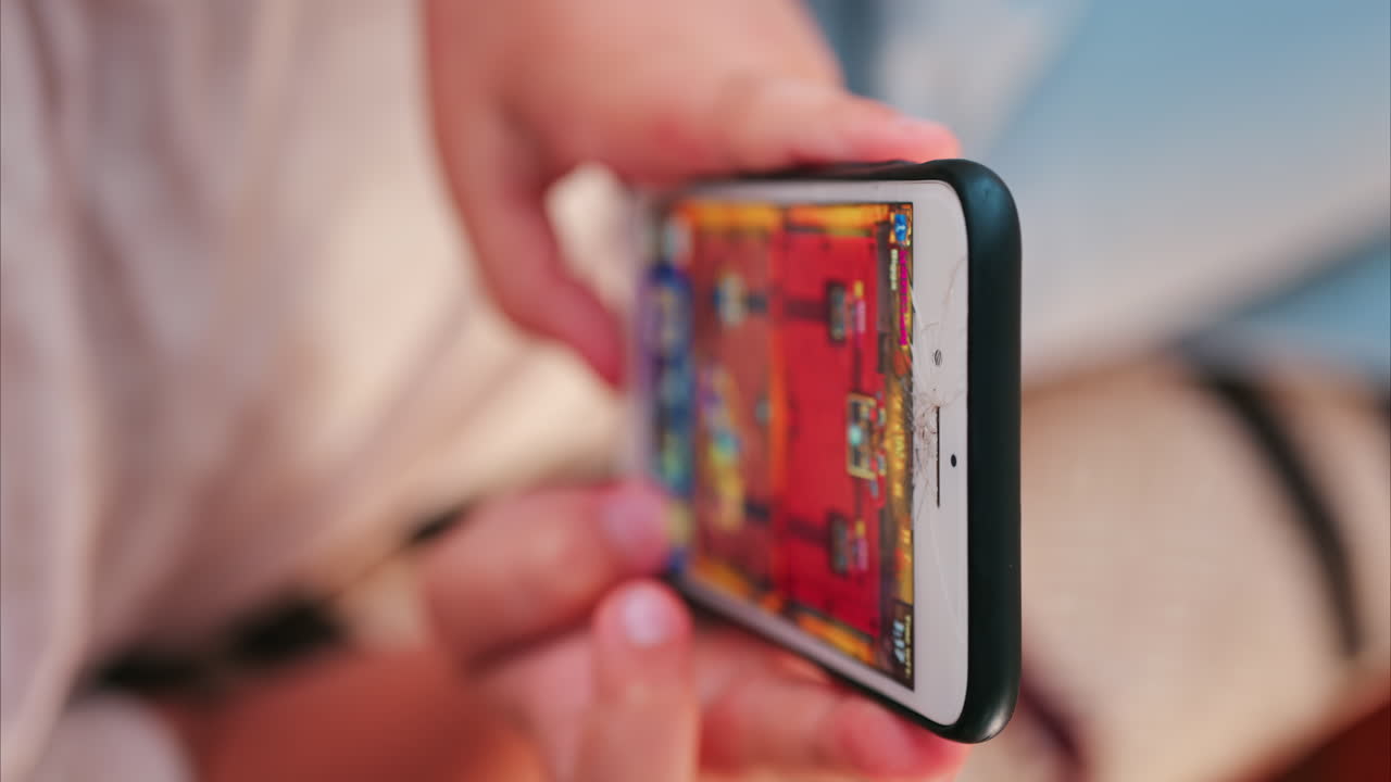 Close up of a smartphone with a cracked screen while a child plays a colourful mobile game. Vertical