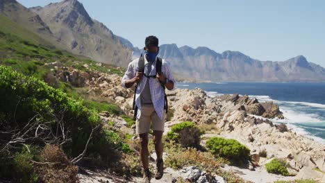 African-american-man-wearing-face-mask-exercising-outdoors-hiking-in-countryside-on-a-mountain