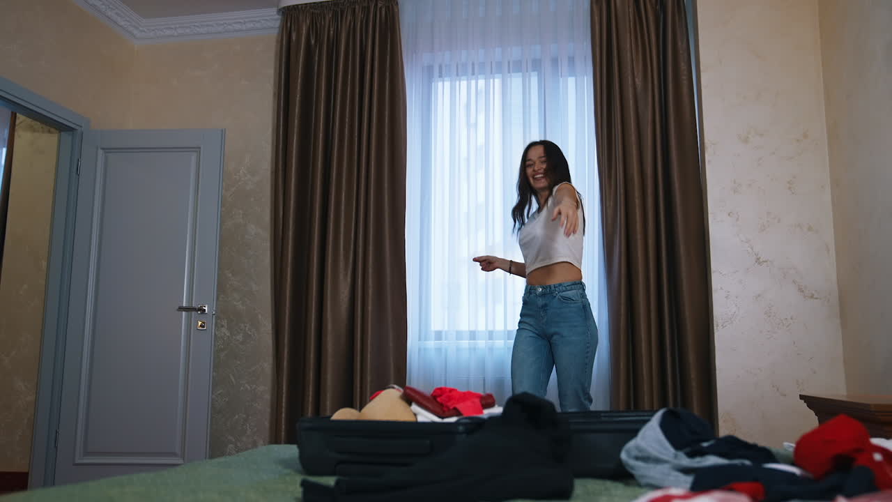 Cheerful woman packing suitcase. Happy girl dancing and putting stuff into a luggage before a journey. Beautiful woman in comfortable room hotel.