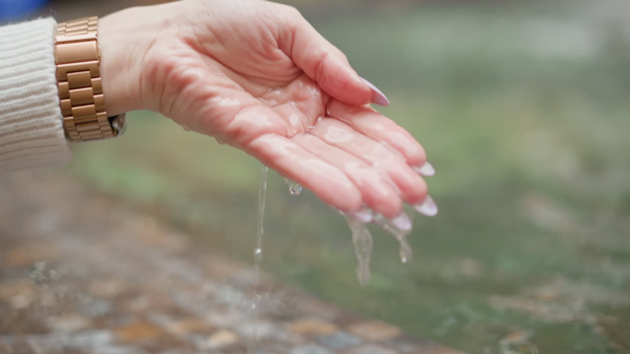 lady hands wearing wristwatch scoop water from mosaic fountain, letting clear droplets spill over fingertips, blurred background jets shimmering under soft mall lighting, and calm ambiance