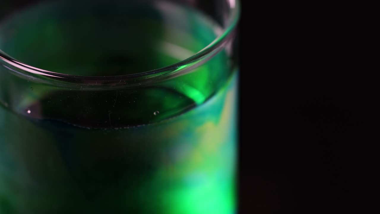 Green liquid disperses in clear water inside a glass, illuminated by dramatic studio lighting