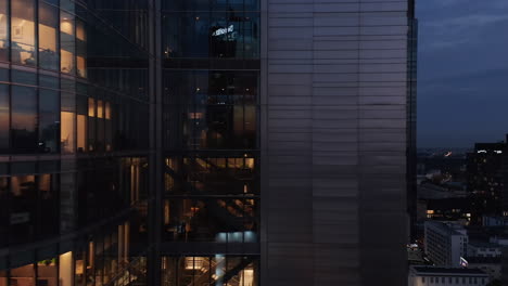 Tight-slide-along-glossy-facade-of-modern-skyscraper.-Downtown-buildings-in-evening-in-background.-Warsaw,-Poland