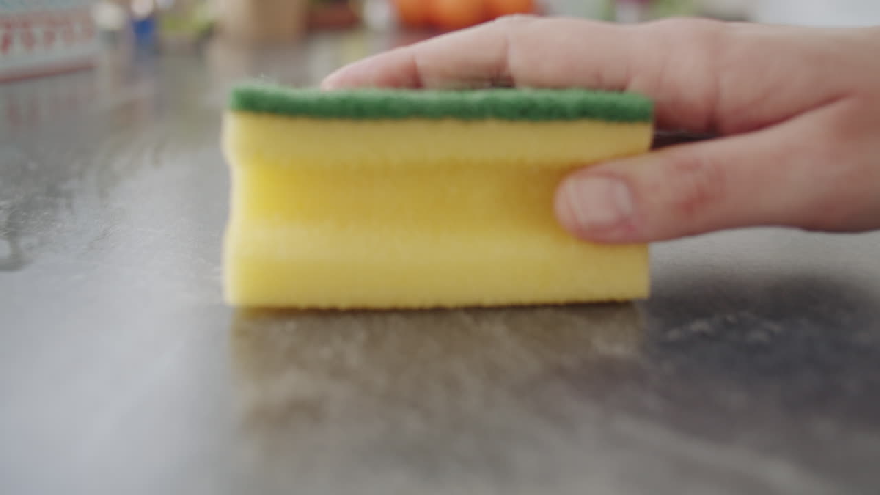 Close up shot of a caucasian hand grabbing a yellow sponge and wiping over a dark kitchen counter. Net with oranges in the background . Camera dollies back. Slow motion shot