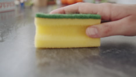 close up shot of a caucasian hand grabbing a yellow sponge and wiping over a dark kitchen counter. net with oranges in the background . camera dollies back. slow motion shot