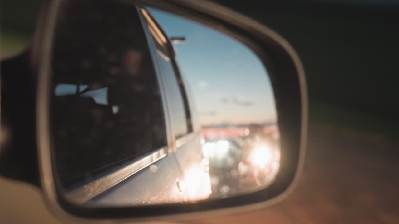 Primo piano di un'auto in movimento con lo specchietto laterale che riflette i fari di un veicolo che si avvicina da dietro su una strada di sera, che mostra la prospettiva del conducente con il riflesso sfocato dei semafori