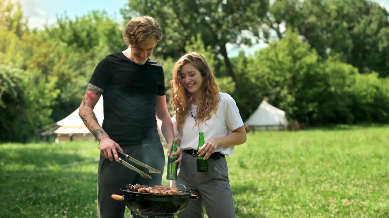 Young couple frying meat on the grill and drinking beer, laughing. Greenery around. Glamping. Slow motion