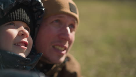 a close-up of a father and his son smiling and looking upwards with joy, the child is dressed in a shiny black jacket and beanie, while the father wears a brown jacket and a beanie