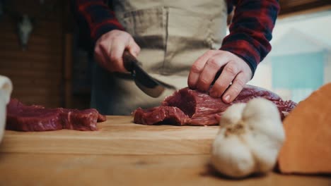 una persona cortando carne roja fresca en una mesa de madera, con ajo y pan en el foco