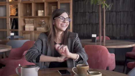 Beautiful-young-woman-in-a-glasses-and-smart-suit-is-sitting-in-a-cafe,-turning-the-music-on-her-phone-on,-swaying-and-dancing