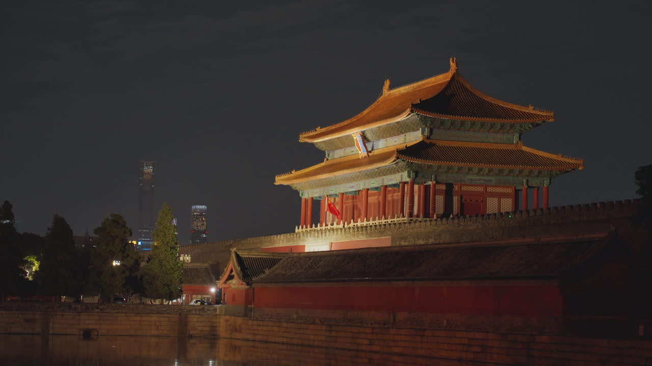 Padiglione cinese illuminato di notte vicino al fossato, tetto dorato e mura rosse che brillano contro il cielo scuro, luci cittadine discrete sullo sfondo, riflessi calmi nell'acqua, architettura imperiale storica immortalata.