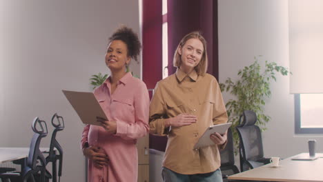 two happy pregnant woman posing at camera while touching her bellies and holding documents and tablet in the office