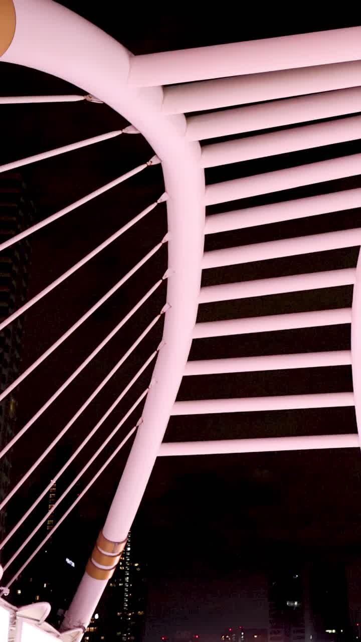 Camera tilts down illuminated bridge structure, revealing pedestrians and cityscape at night in Bangkok