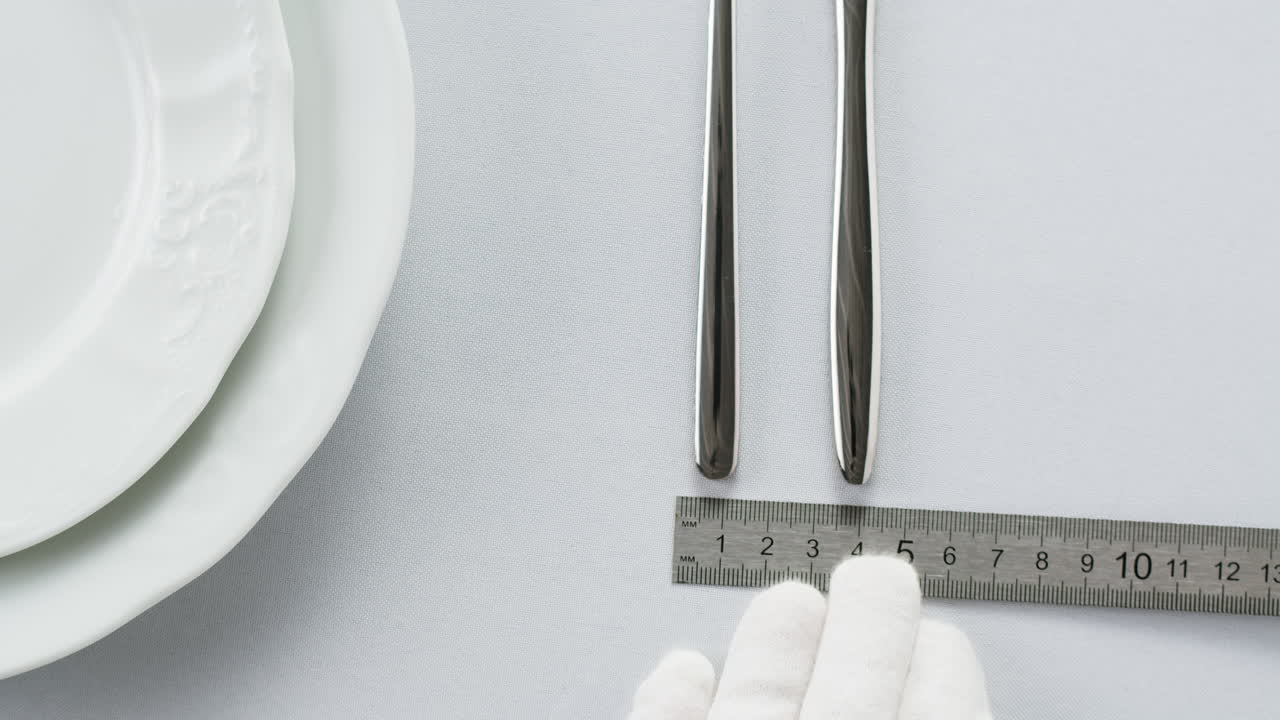 Professional table setting with white gloves. Plates, fork, knife, and glass arranged on a white table.