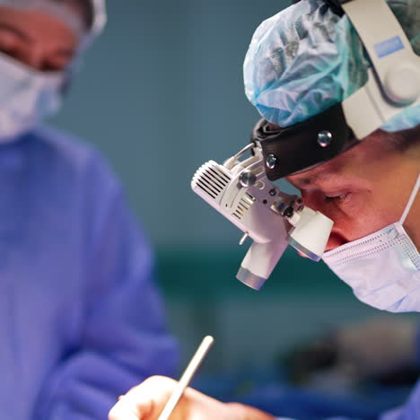 mid-aged caucasian surgeon in cap, mask and binocular loupe device uses metal tool. side view close up portrait of a neurosurgeon at work.