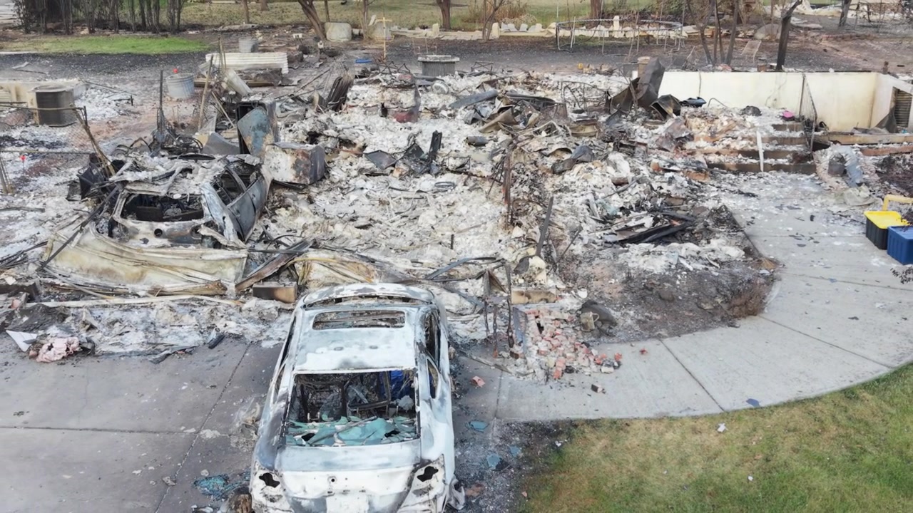 Danni dell'incendio di Spokane che mostrano una ripresa aerea bassa e in movimento all'indietro sopra un'auto bruciata e un lotto residenziale carbonizzato, con detriti e distruzione causata dal fuoco dopo l'incendio del 2026.