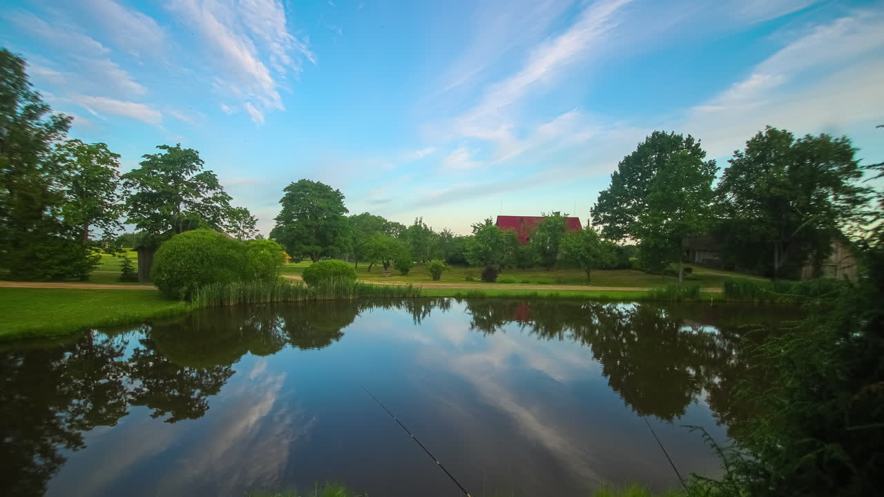 나무, 잔디, 뒷면에 빨간 지붕이 있는 집과 함께 조용하고 반사되는 호수 위의 일출의 영화적 타임 스