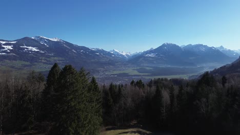 Drone-rise-up-shot-of-green-field-with-small-huts-in-foreground-and-cityscape-and-snowy-mountains-in-background,-Aerial-view-over-Nenzing-Austria