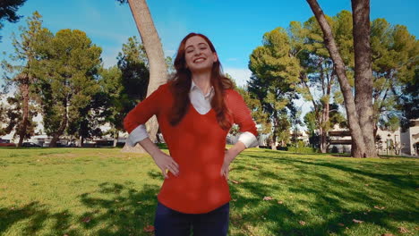 una feliz, atractiva, joven, mujer caucásica en el parque, pone sus manos en sus caderas y sonríe con gran confianza