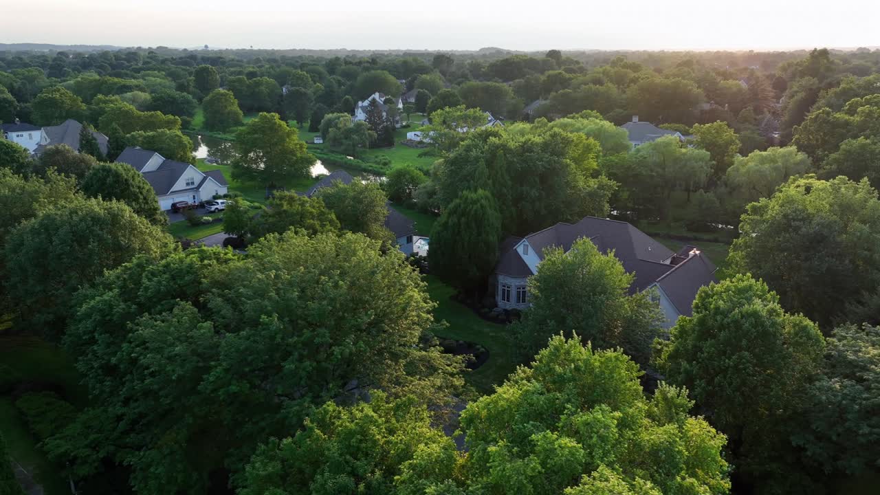 Quiet sunrise in well-maintained suburb district of town. River flowing between single family houses and one family homes in America. Aerial orbit wide shot. Green trees in summer season