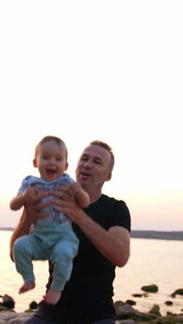 lovely happy baby boy is tossed in the air by his dad. father and son having good time near the river at sunset. vertical video
