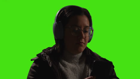 close up of woman wearing wireless headphones streaming music from mobile phone against green screen with low key lighting