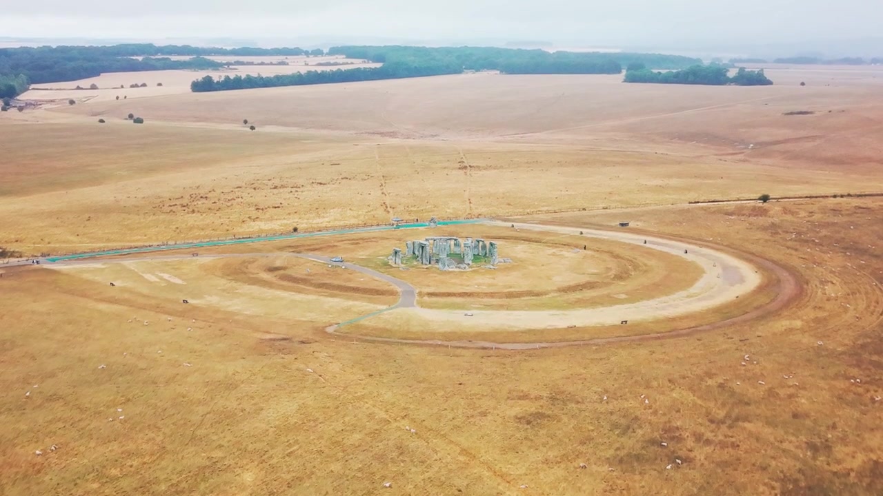 Bredt luftpanorama viser Stonehenge isoleret i udstrakte gyldne marker og bølgende landskab under en bleg, diset himmel.