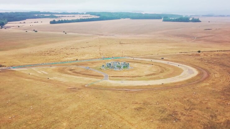 Wide aerial panorama shows Stonehenge isolated within expansive golden fields and rolling rural countryside beneath a pale hazy sky