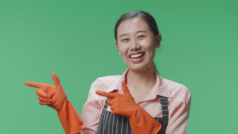 primer plano de una ama de casa asiática con un delantal sonriendo y señalando al lado mientras está de pie en el estudio de pantalla verde