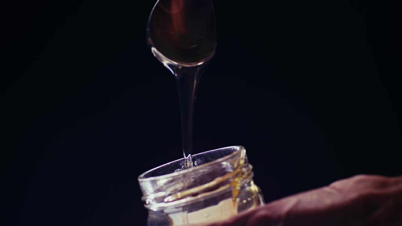 Chef's hand elegantly putting spoon of honey in a glass jar against a black background. Slow motion shot with professional studio lighting