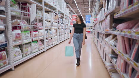 young lady in black top and jeans walking through supermarket aisle near textile products, focusing on goods displayed on shelves while holding shopping bags, with another person standing behind