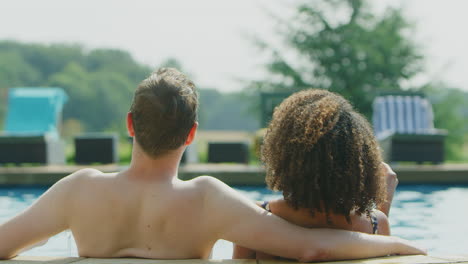 Vista-Trasera-De-Una-Pareja-Amorosa-Relajándose-En-La-Piscina-De-Vacaciones-Celebrando-Bebiendo-Champán