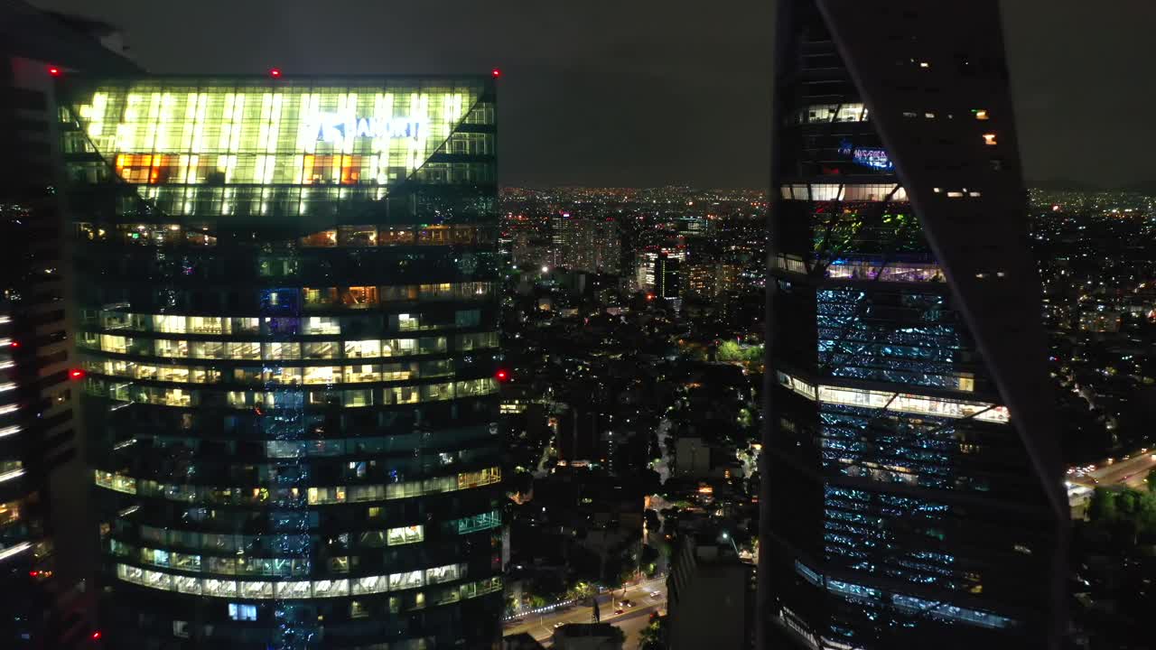 Exhilarating high aerial flight through narrow opening between tall torre mayor tower skyscraper and reforma glass building lit with PRIDE colors at night, Mexico city, drone going away