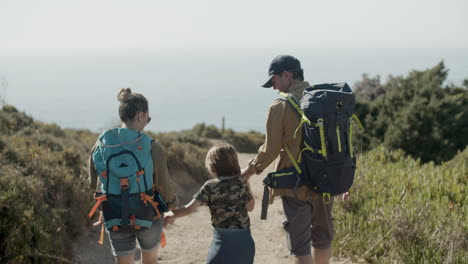 Parents-carrying-backpacks,-holding-child's-hands-and-walking-along-dirt-road