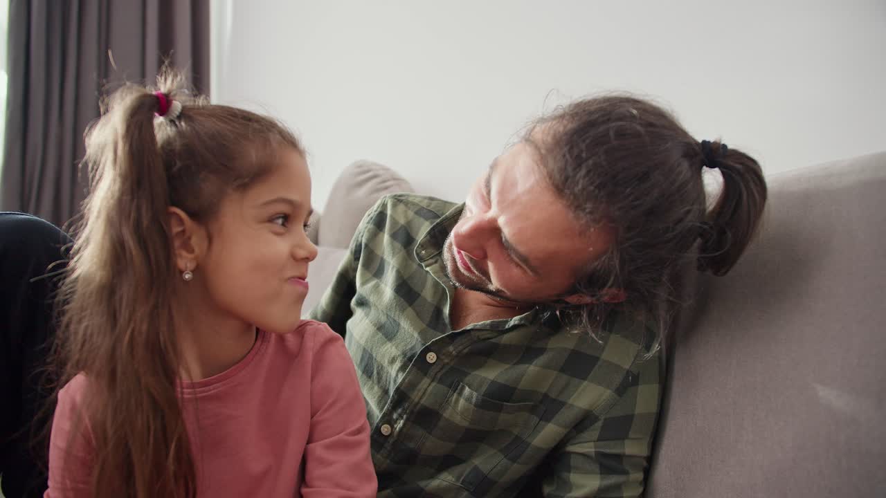 un hombre moreno con rastro en una camisa verde a cuadros se sienta en el sofá y se comunica con su pequeña hija morena en una chaqueta rosa en un hogar moderno. feliz hombre moreno padre soltero se comunica con sus hijas pequeñas mientras está sentado en un sofá gris en un apartamento moderno