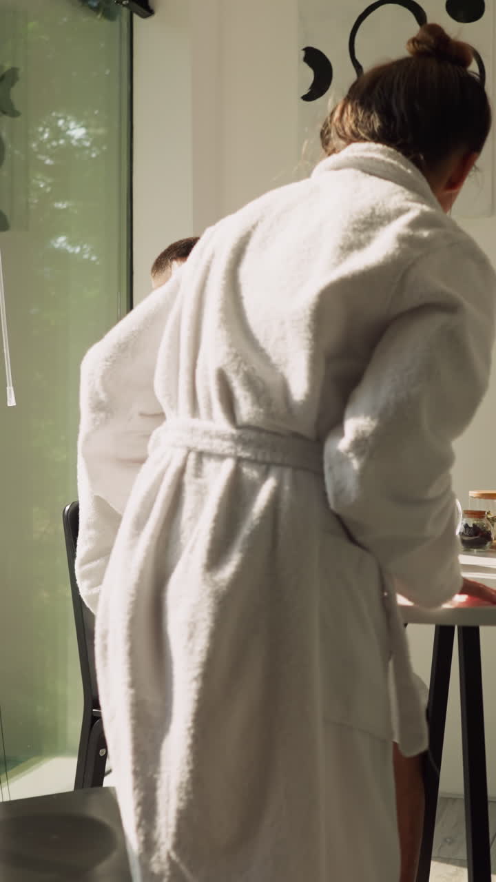 Woman comes to man sitting at table. Young sleepy wife in white robe sits down on chair by husband to drink tea in morning. Happy couple lifestyle