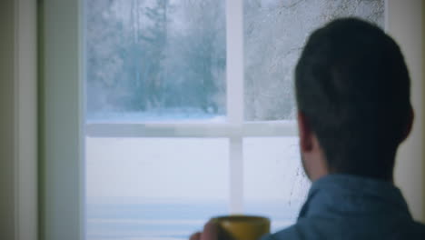 man in blue shirt enjoying hot drink, gazing through frosty window, feeling cozy indoors