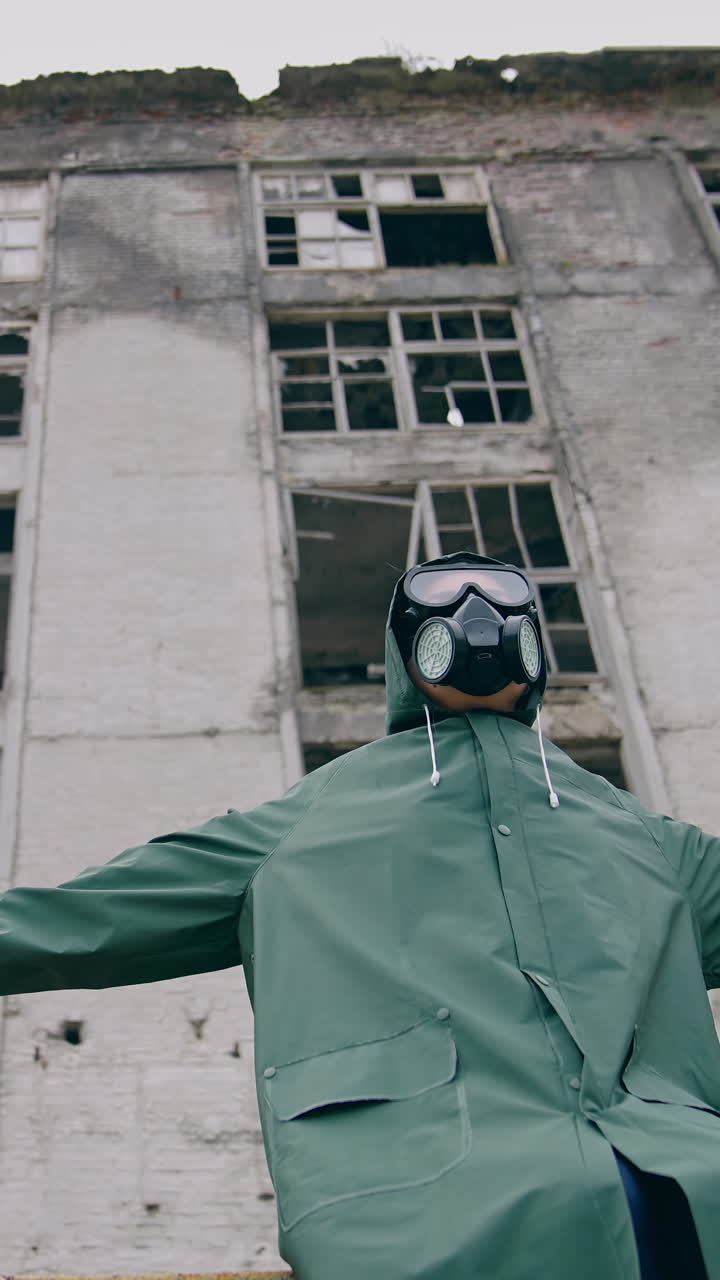 Man with gas mask in ruined place. Human in protective suit and respirator sitting outdoors with outstretched arms on the background of destroyed building. Radioactive zone. Vertical video