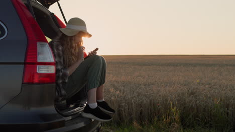 Una-Adolescente-Sentada-En-El-Maletero-De-Un-Auto,-Usa-Un-Teléfono-Inteligente.-Con-El-Telón-De-Fondo-De-Un-Paisaje-Rural-Donde-Se-Pone-El-Sol.-Vídeo-4k