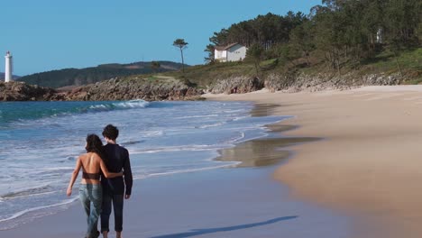 view behind tourist couple strolling barefoot at serene beach with lighthouse in distance. muxia, spain. static shot