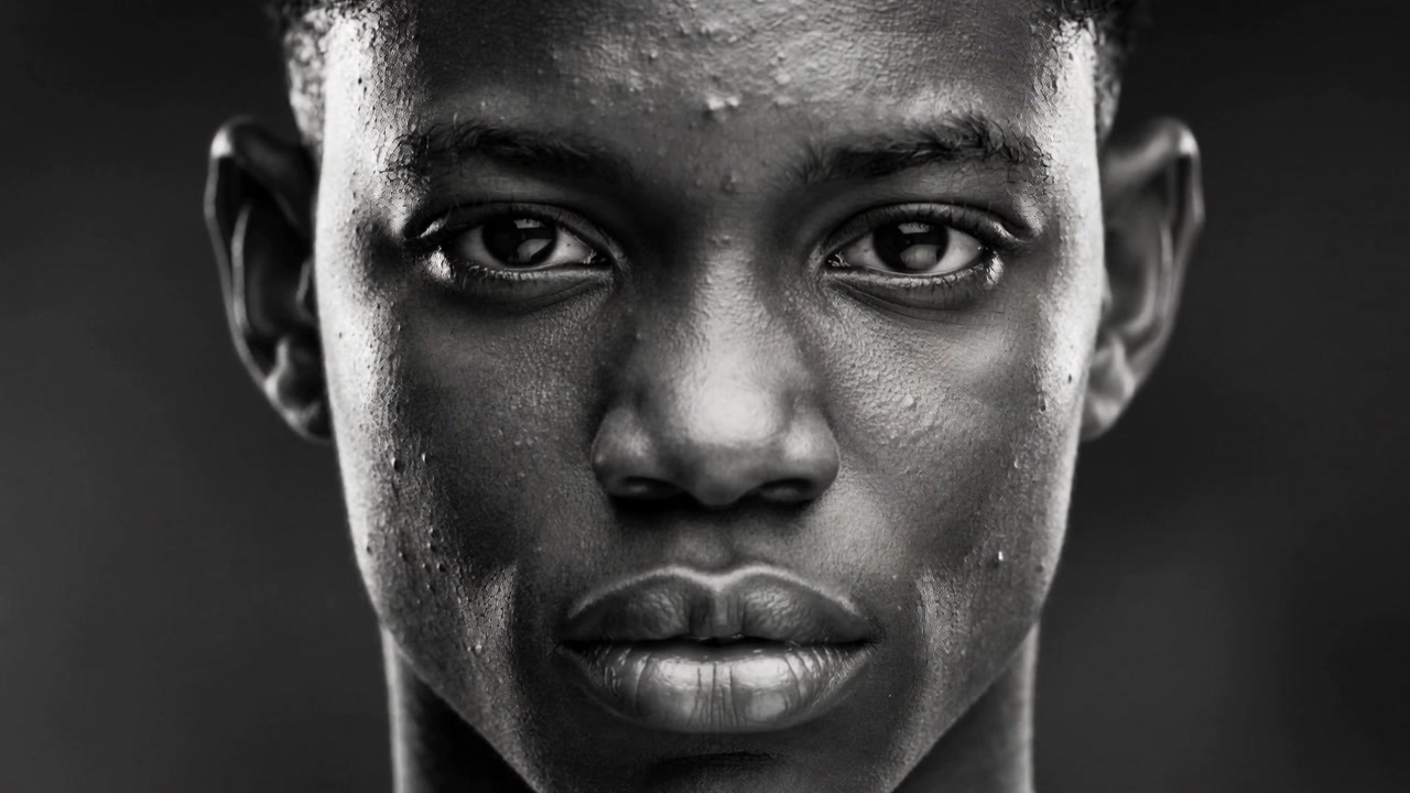 Adolescente negro de frente, mantendo o olhar enquanto a câmera aproxima em um retrato de estúdio, com luzes mostrando umidade.