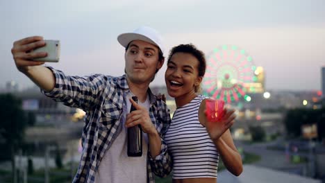 cheerful multiethnic couple caucasian young man and mixed race girl are taking selfie on rooftop using smartphone, laughing and holding drinks.