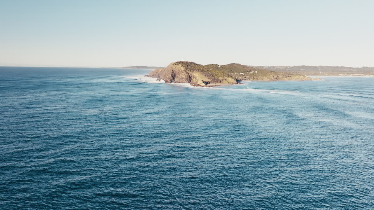 Superficie dell'oceano aperto con dolci increspature e costa distante sotto un cielo limpido, dolly aereo verso le scogliere di Byron Bay, Australia