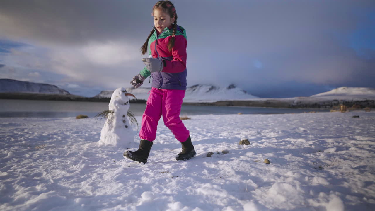 Kid in vibrant clothes kneeling in snow creating snowman during family trip